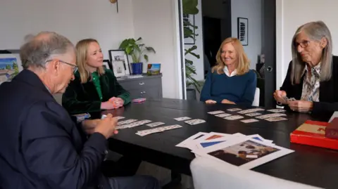 Maxine Collins/BBC (From left to right) Side profile of Bill Gates holding cards, Katie Razzall smiling, with Bill's sisters Libby and Kristi, who is playing cards 