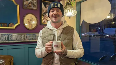 Chris Taylor, owner of Gran T's in Manchester, smiling while holding a branded coffee mug