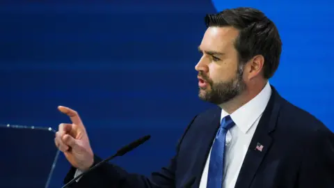 Reuters JD Vance, wearing a dark blue suit and blue tie with a US flag lapel badge, points his finger while giving a speech at the Munich Security Conference.