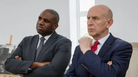 Reuters David Lammy and John Healey wear suits and have their arms crossed as they stand and look forward. Lammy is wearing a grey suit and tie while Healy is wearing a blue suit and red tie.