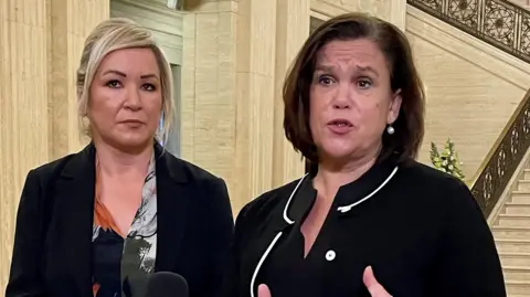 PA Media Michelle O'Neill has blond hair tied in a ponytail and is wearing a black suit jacket and patterned blouse. Mary Lou McDonald has short dark hair and is talking while wearing a black suit jacket