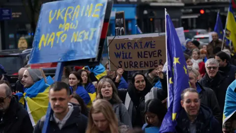 PA Media Protesters march in London, some wrapped in Ukrainian flags. One is holding an EU flag while two posters are legible, one reading: Ukraine now Nato next