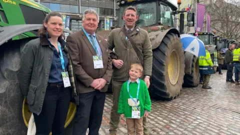 Malcolm Prior/BBC Hazel and Tom Church with Martin Towler, and five-year-old Bertie, standing by a line of tractors in a wet cobbled street. Other farmers stand in the background in hi-viz jackets