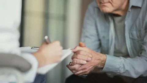 Getty Images A doctor writes on a clipboard as a patient, sitting with hands clasped, listens