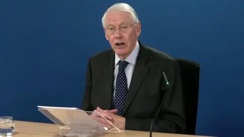 PA Media Image shows Sir Martin Moore-Bick, a Caucasian man with white hair and glasses, wearing a black suite with striped shirt and a blue tie with white spots, sitting at a desk against a blue backdrop