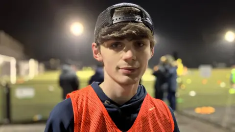 BBC / Kate McGough Luke looks into the camera in the centre of the image, with blurred astro football pitches lit by floodlights in the background. He has a backwards cap on with hair down over his forehead,  and he is wearing an orange bib over his hoodie.