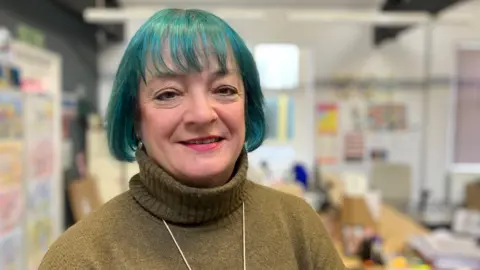 BBC / Kate McGough Gill Bainbridge looks into the camera, smiling. She is wearing a yellow-green turtle neck jumper and has a short bob, dyed blue.