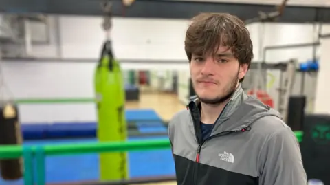 BBC / Dan Nelson Niall looks into the camera. He has medium-length brown hair down over his forehead and is wearing a grey North Face zipped-up jacket. In the blurred background we can see he is in a boxing gym.