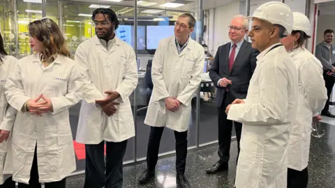 Sir Sadiq Khan is wearing a white hard hat and white lab coat, stood amongst oa group of men and women also wearing whit lab coats with the wording 'Imperial' in blue. One man with white hair and glasses is wearing a navy suit and red tie. 