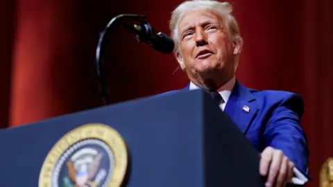 Reuters Donald Trump speaks at a podium. He has grey hair and is looking upwards to his left and is wearing a blue suit and white shirt