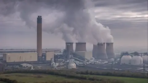 Getty Images Drax power station. The photo shows smoke billowing out of six stout chimneys. The sky is cloudy and smoky. There are trees and grass in the foreground.