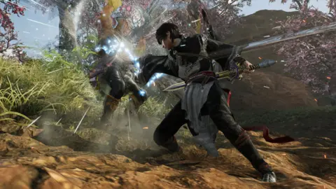 Koei Tecmo A character attacks another, causing him to fly backwards with light refracting off his body. They are both dressed in feudal era Chinese clothes and wielding swords. Behind them, a lush green scene with cherry blossom trees.