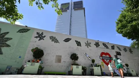 Getty Images A woman and girl look up at Grenfell Tower from in front of a wall covered in written tributes