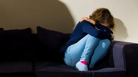 PA Media A woman covers her face with her arms as she cowers on the corner of a sofa