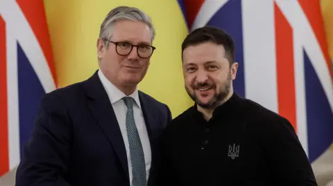 Reuters Sir Keir Starmer, with combed-back grey hair and wearing black glasses, a dark blue suit, white shirt and blue patterned tie, stands next to a smiling Volodymyr Zelensky, who is shorter, has short black hair and a beard with a dark shirt.