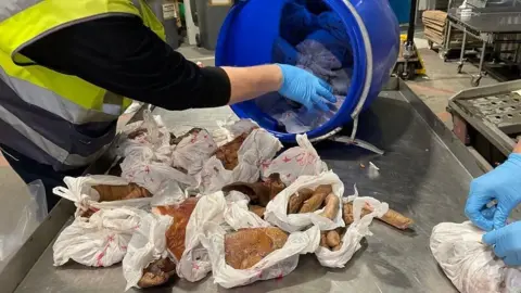 Dover Port Health Authority A person wearing a high-vis jacket and blue plastic gloves pulls sausage meat wrapped in white plastic bags out of a blue bucket