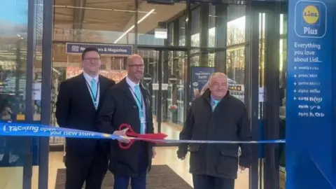 Facebook/David Thomas The three Reform councillors all wearing black jackets, white shirts and pale blue ties. Two are bald, while the third has short dark hair. The one in the middle is holding a pair of giant red scissors as they stand behind a Lidl branded ribbon which has the words 'new store' on it, with the glass doors of Lidl in Cwmbran behind them.