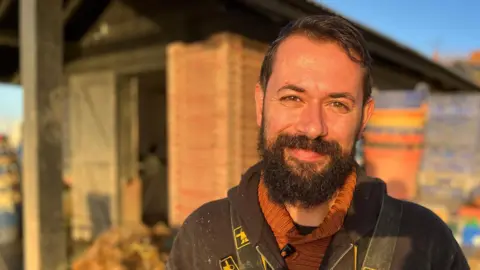 BBC Tom Haward, a man in his early forties with a thick beard, smiling directly at the camera. He is standing in front of his oyster shed on Mersea Island in Essex.