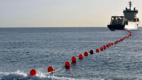 Getty Images A boat in the distance with a cable connected by orange bollards floating on the sea.