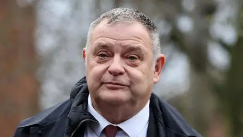 Reuters Mike Amesbury arrives at Chester Magistrates' Court ahead of his sentencing hearing on Monday. He wears a dark red tie, white shirt and a dark coat.