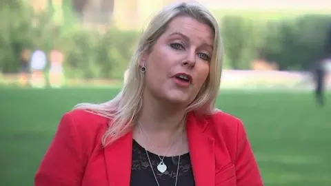 BBC Mims Davies, who has long blonde hair, wearing a red jacket and black top as well as a silver necklace and earrings. Behind her is a green lawn.