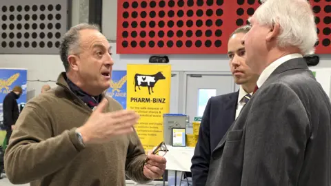 Paul Moseley/BBC Mark Means is challenging Daniel Zeichner. Mr Means is gesticulating with his hand as he speaks - he's wearing a brown jumper on top of a blue shirt and tie. We can't see Zeichner's face from this angle - but he's wearing a grey suit jacket, white shirt and red tie.