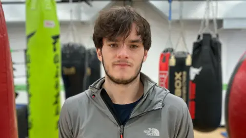 BBC / Dan Nelson Niall looks into the camera. He has medium-length brown hair down over his forehead and is wearing a grey North Face zipped-up jacket. In the blurred background we can see he is in a boxing gym.