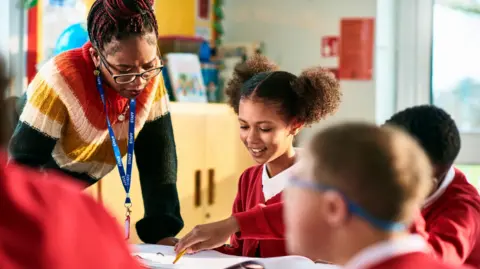 Getty Images A teacher wearing a brightly coloured knitted jumper leans over a table in a classroom where children in red uniform cardigans are doing their work. In focus is a girl who is smiling down at the paper in front of her.