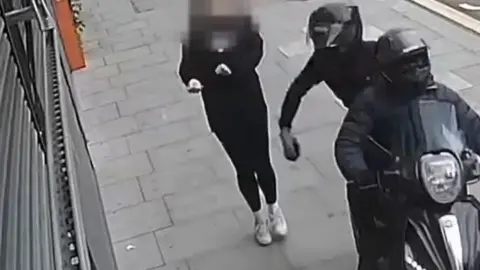 Metropolitan Police/PA Wire Two people riding a moped dressed in black and wearing helmets snatch a phone from a woman, whose face is blurred in the image, on the pavement