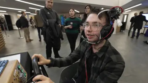 BBC A man wearing glasses and a green check shirt smiles as he wear a set of headphones with wires and tubes attached to deliver smells in front of the user's nose. He has his hands on a steering wheel playing an arcade game.