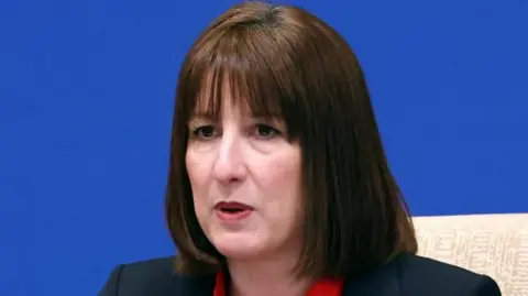 Reuters A headshot of Rachel Reeves, with shoulder-length brown hair, wearing a suit jacket and red top, against a blue background at the China-UK Financial Services Summit in Beijing
