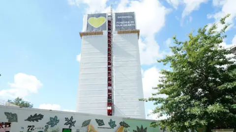 PA Media Image shows Grenfell Tower and the a wall full of memorials and messages on a sunny day, with blue sky and clouds