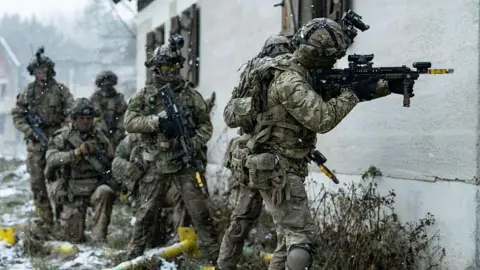 MOD British Army officer cadets from the Royal Military Academy prepare to assault a building during an exercise in Germany. They wear combat uniforms and wield guns as snow falls around them.