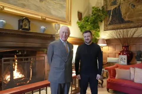 Reuters King Charles wearing a grey suit standing next to Volodymyr Zelensky in a wood-panelled sitting room with a fireplace and red sofa with striped cushions.