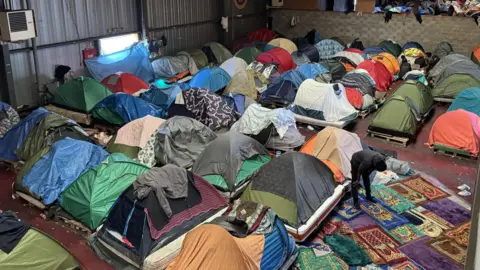 Jack Valpy/BBC Several tents inside a warehouse. Prayer mats form a makeshift area for those seeking to pray. 