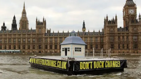 Oli Scarff/Getty Images A mock giant duckhouse on a floating platform is towed along the river Thames, with the Houses of Parliament visible in the background. There are black banners with writing in yellow capital font on the sides of the platform, one listing the campaign group website www.voteforachange.co.uk and another with the slogan 