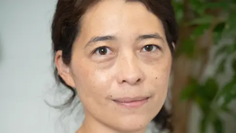 Lee Townsend Headshot of Akiko Hart looking directly at the camera. Her eyes are brown, her black hair is tied behind her head and she has pursed lips. Over her left ear, there is an out-of-focus plant in the background.