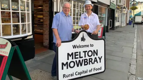 Matthew O'Callaghan (left) wearing a blue shirt next to a man from Dickinson & Morris pork pie bakery wearing a white shirt and hat. They are stood holding a white sign saying 