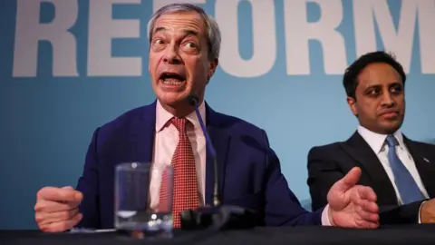 Getty Images Nigel Farage speaking into a microphone positioned on a table, next to Zia Yusuf who sits further back and appears to be listening 