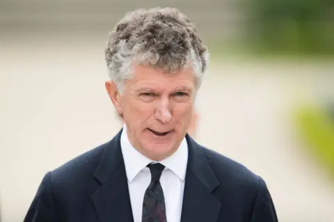 Getty Images Mid-shot of Jonathan Powell with curly ash hair wearing a black suit, white shirt and black tie.