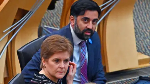 Getty Images Nicola Sturgeon sitting in front of Humza Yousaf are their seats in the Scottish Parliament chamber. Sturgeon is touching her left cheek with her right hand and is wearing a black shirt white white hoops. Yousaf is looking in the direction of the camera and is wearing a blue suit, white tie and purple and blue tartan tie. 