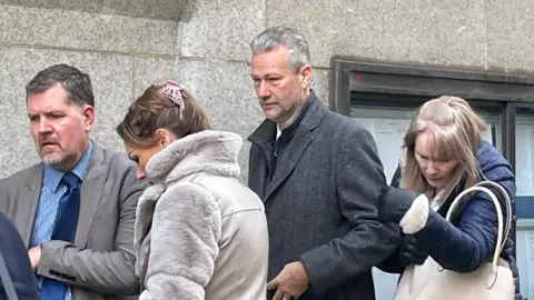 A man with short grey hair, wearing a smart grey coat, stands in a queue, looking down. A blonde woman in a blue coat with a cream handbag is behind him, and a man in a grey suit and striped shirt alongside a brunette woman in a furry grey coat are stood in front of him. None of them are looking at the camera.