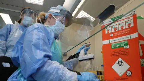 Getty Images Two medical technicians are wearing PPE at Queen Elizabeth University Hospital in Glasgow on April 22, 2020. Both are wearing blue, plastic aprons, surgical masks and visors. There is a sign behind them which says 