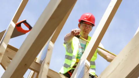 Getty Images A builder in fluorescent yellow vest and a red hard hat measures struts for a roof. He is standing on the struts and photographed from below. 