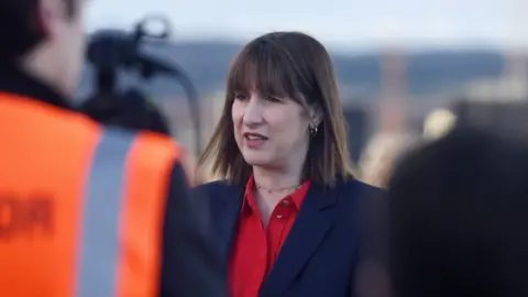 PA Media Chancellor Rachel Reeves speaks to reporters, wearing a scarlet open necked shirt, a navy suit jacket and a gold choker chain, with small gold hoop earrings. 