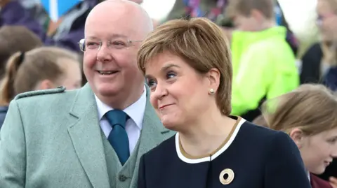 PA Media Peter Murrell and Nicola Sturgeon before at an event their separation. Murrell has a bald head and is wearing thin-framed glasses, a green jacket and waistcoat with a white shirt and blue tie. Sturgeon is smiling and is wearing a blue dress. There are children in the background