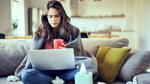 Getty Images A sick worker sits at home on their laptop wrapped in a blanket surrounded by used tissues