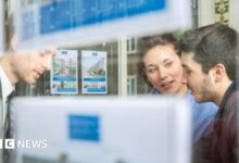 A man and woman talk to an estate agent in their office, pictured through the window.