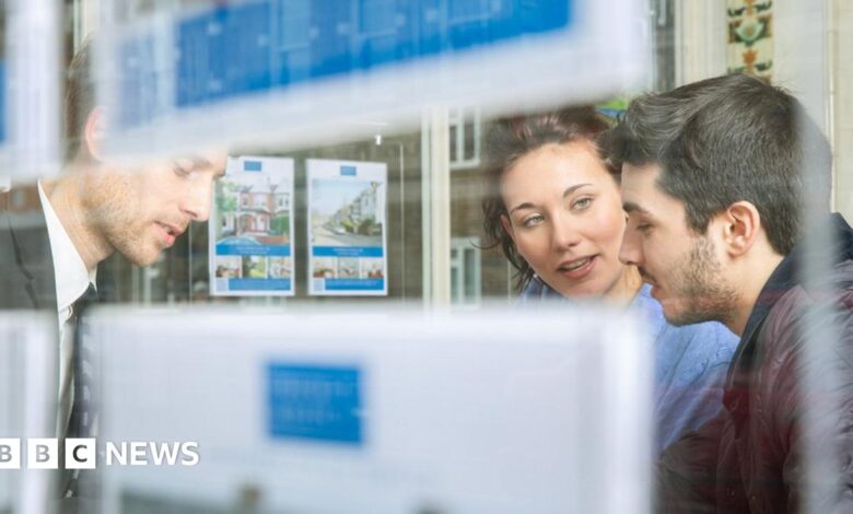 A man and woman talk to an estate agent in their office, pictured through the window.