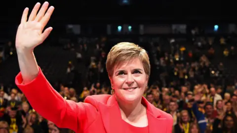 Getty Images A woman, photographed from the chest up, looking up and waving with her right hand. She has short brown hair, combed to the side, and is wearing a red top and matching red jacket. A large crowd of people are visible in the background in the image, which was taken in an auditorium. 